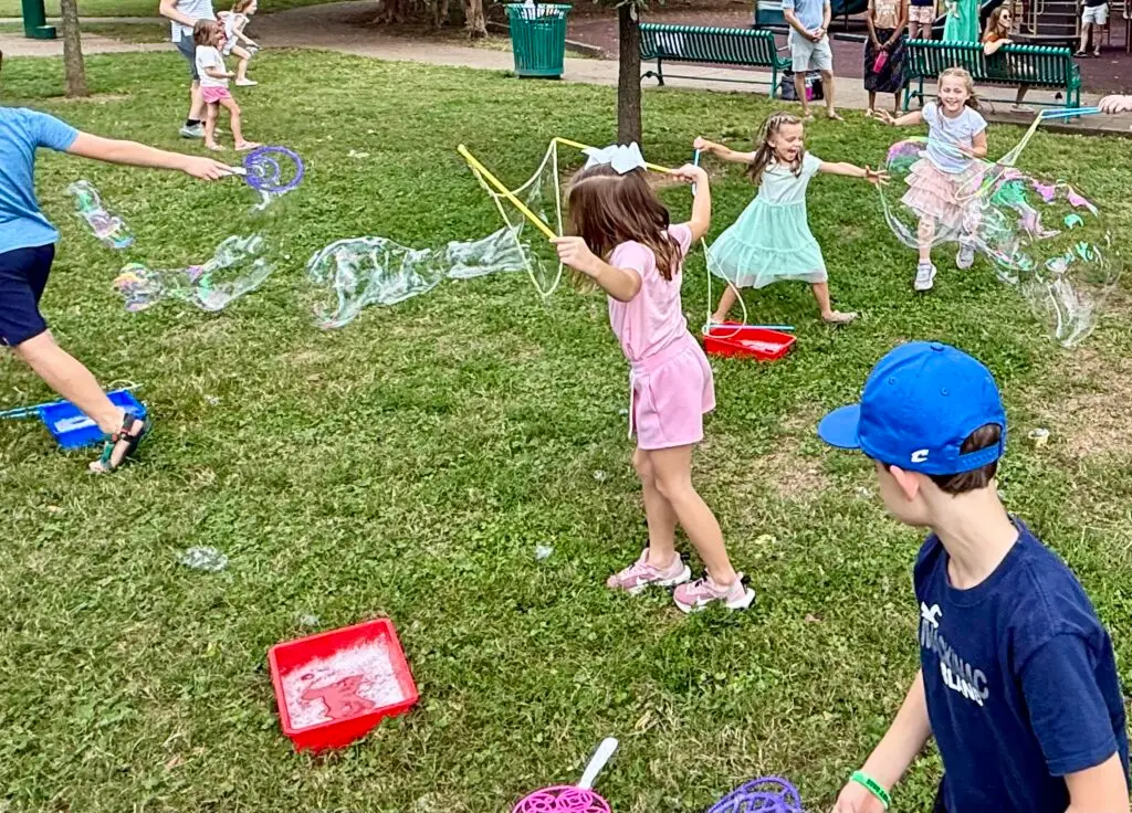 Kids making giant soap bubbles with wands and trays at a park bubble party