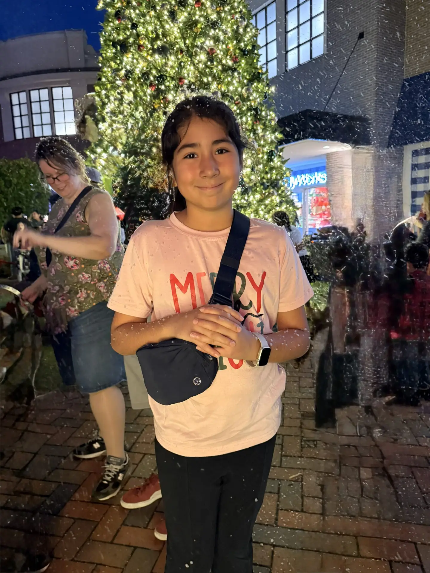 A smiling child stands in front of a decorated, lit christmas tree at night, wearing a merry vibes shirt and crossbody bag, with people and festive lights visible in the background. | hilltop foam parties