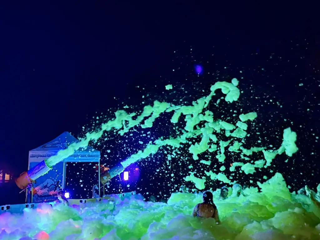 People enjoy a nighttime foam party outdoors, with neon-green foam spraying from cannons and glowing under colorful lights. A white tent and purple lighting are visible in the background. | Hilltop Foam Parties