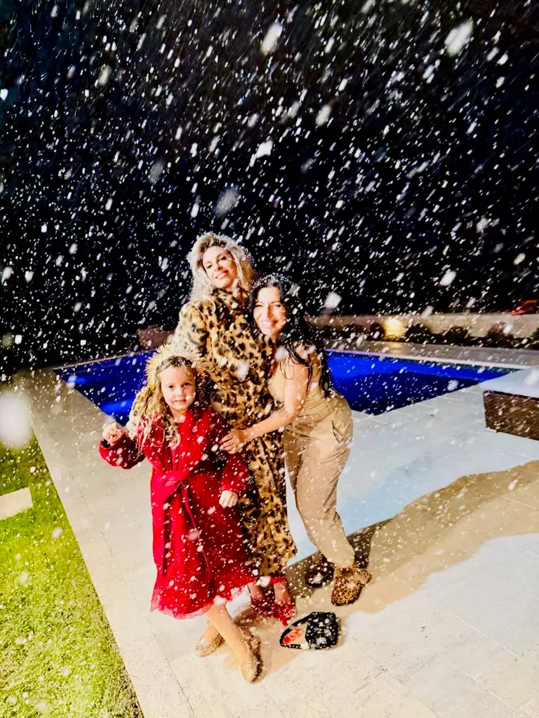 Three people stand smiling in falling snow by a lit outdoor swimming pool at night, enjoying the snowy moment made possible by a snow machine rental—one child in a red coat, an adult in leopard print, and another in beige pose together. | hilltop foam parties