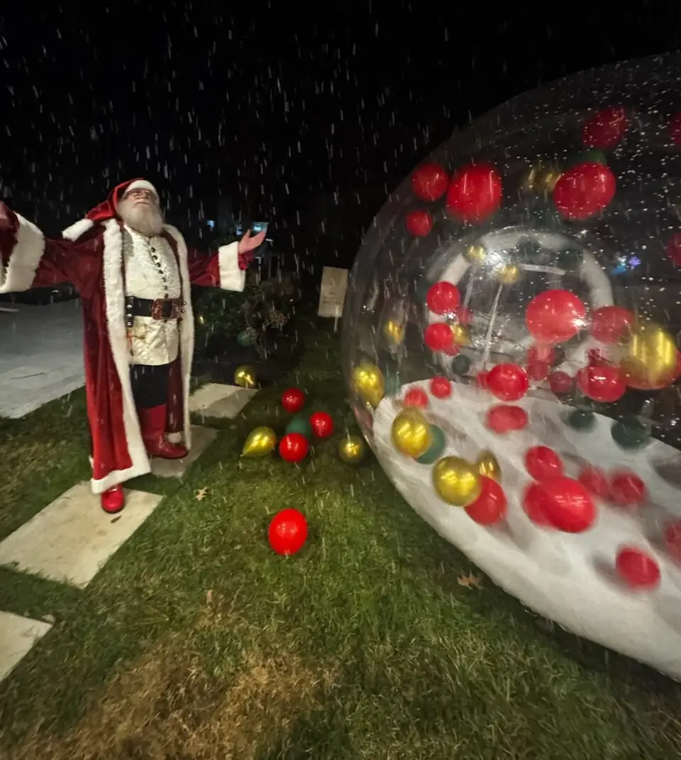 A person dressed as santa claus stands on a stone path with arms raised beside a large transparent snow globe filled with red and gold ornaments and artificial snow, as snowfall from a snow machine rental adds magic to the winter night. | hilltop foam parties