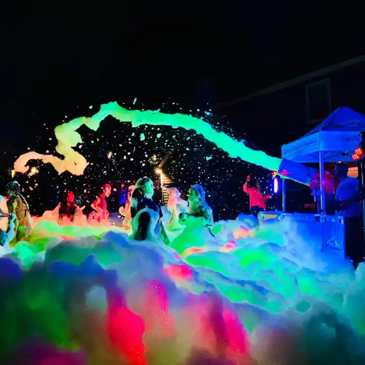 People enjoying a nighttime foam party with colorful lights illuminating thick foam and bubbles, creating a vibrant, festive atmosphere outdoors. | hilltop foam parties
