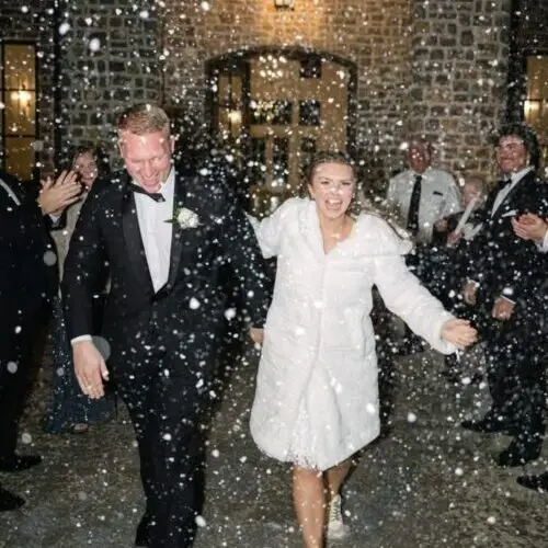 A joyful couple in formal attire, the woman wearing a white coat, walk hand in hand through falling snow, surrounded by applauding guests outside a brick building at night. | hilltop foam parties