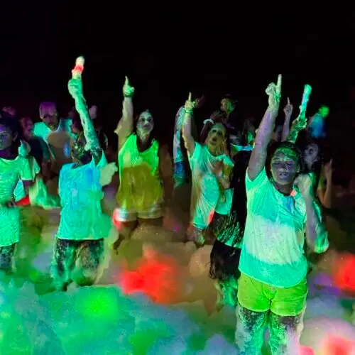 A group of people in white clothes dancing and cheering in colorful neon lighting and foam at a nighttime foam party. | hilltop foam parties